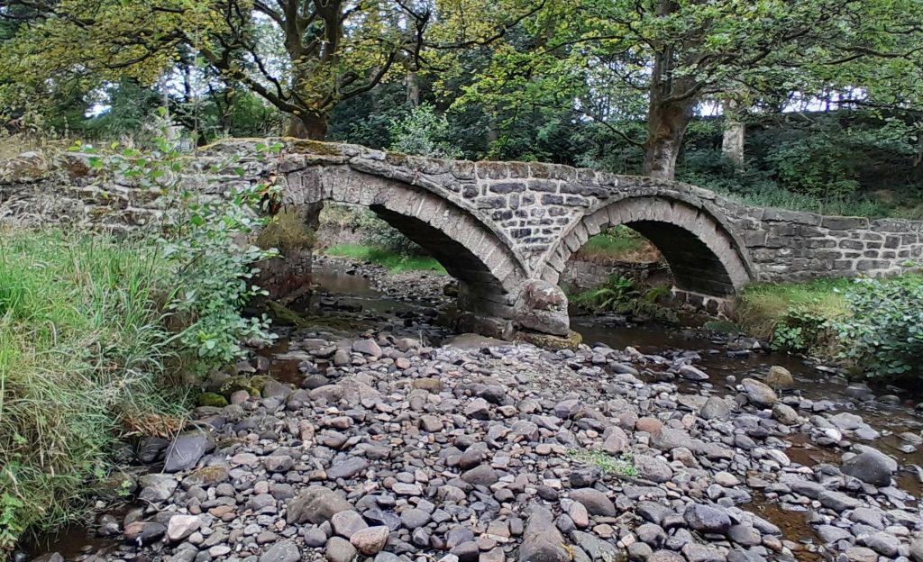 A view of the side of the Packhorse Bridge at Wycoller. Stone bridge with two arches crossing the stream and stoney riverbed, with trees in the background. 