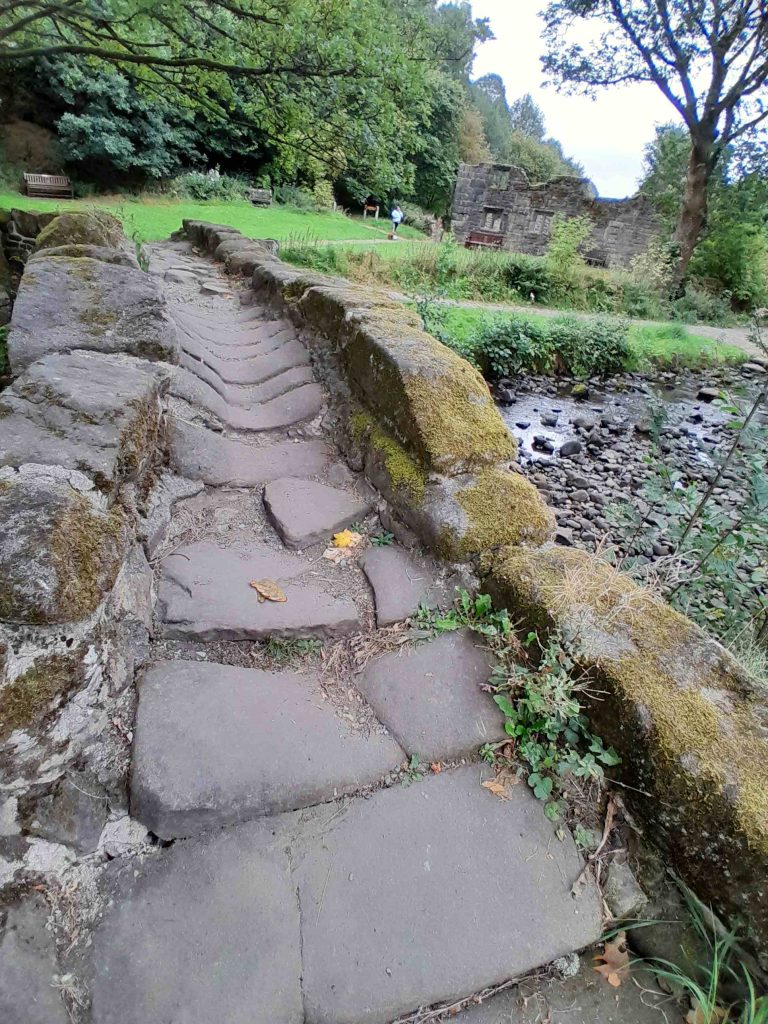 A view of the top of the packhorse bridge at Wycoller, very narrow the stones are uneven and worn down in the middle and very slippy with age. In the background is the ruins of Wycoller Hall.