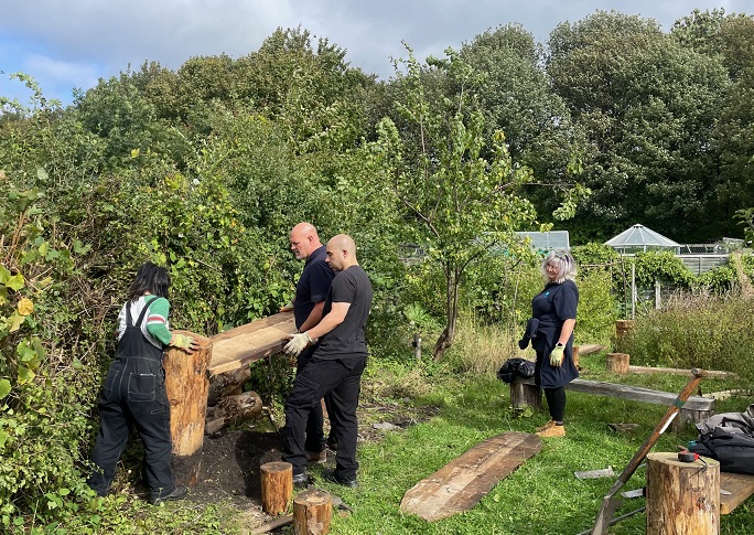 a group of people working together to measure a wooden bench in an allotment