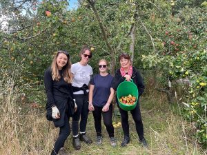 4 women standing under an apple tree. One is carrying a bucket of apples they have collected